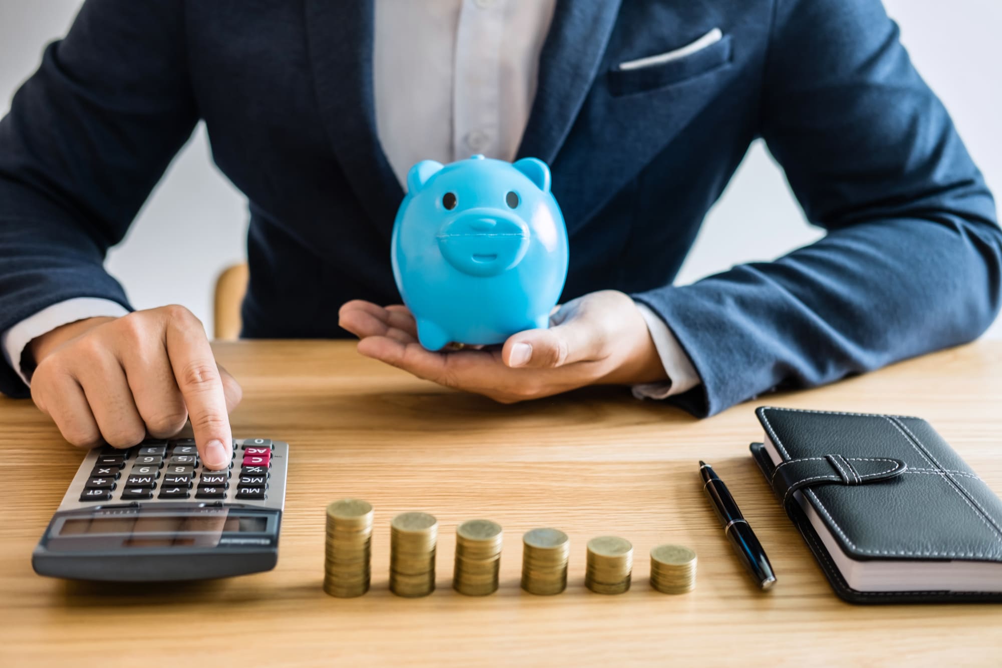 コインが並ぶ前で豚の貯金箱を持ち電卓をたたくビジネスマン